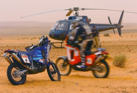Un partecipante passa accanto alla moto di Fabrizio Meoni mentre lo sfortunato pilota italiano viene portato via dall'elicottero sullo sfondo dopo la caduta che l'ha ucciso l'11 gennaio 2005 (AP Photo/Bernard Papon, Presse Sports)